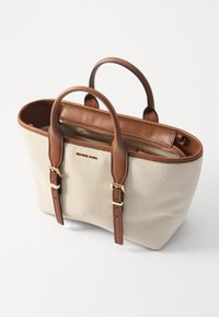 Beige canvas kantokassi, jossa on ruskeat nahkakahvat ja -koristeet. Kassi sisältää vetskalla suljettavan sisätaskun ja kullanvärisen logoyksityiskohdan etupuolella.