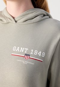 Sweat à capuche gris clair avec texte blanc "GANT 1949" et un petit logo en forme de bouclier avec le drapeau américain au-dessus de rayures rouges et blanches sur la poitrine.