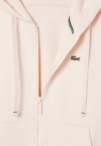 Sudadera con cremallera de color rosa claro hecha de tejido texturizado, con un pequeño logo verde de Lacoste, cremallera blanca y detalles de cordón.
