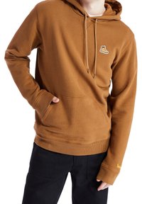 Persona con una sudadera marrón con un pequeño logo de bota en el pecho y pantalones negros, con una mano en el bolsillo delantero.