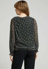 Chemisier noir transparent avec des fils métalliques, présentant des motifs floraux éparpillés et des poignets à volants. La vue de dos met en avant son design texturé.