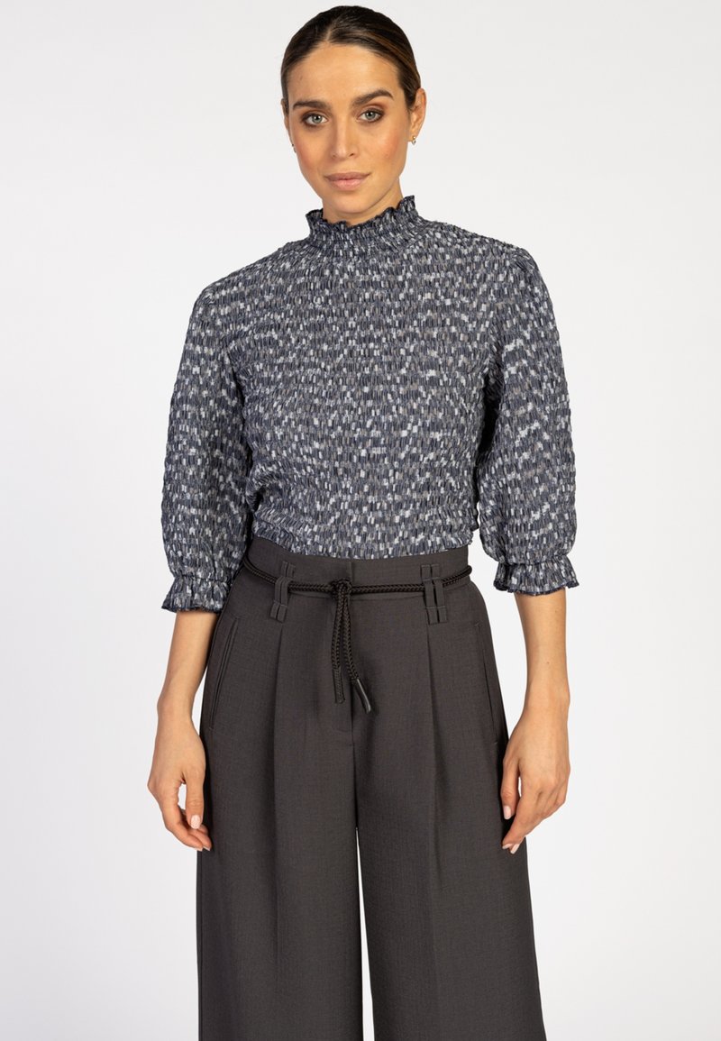 Vrouw in een textuurrijke blouse met hoge hals en driekwartmouwen, gecombineerd met donkere, wijde broek met een dunne riem om de taille.