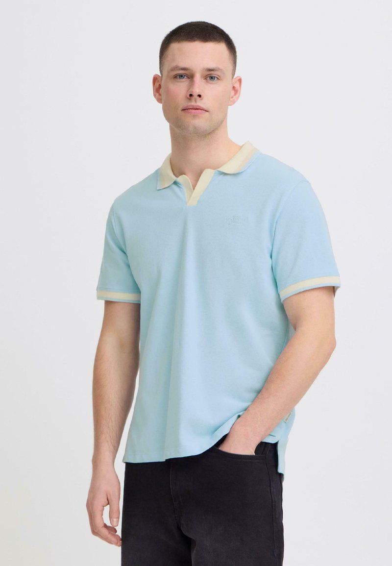 Jeune homme portant un polo bleu clair avec un col beige et un bord de manche, debout avec une main dans la poche de son pantalon noir, devant un fond uni.