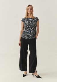 Top senza maniche con un motivo astratto blu e beige, abbinato a pantaloni neri a gamba larga con slitte frontali e scarpe nere a punta.