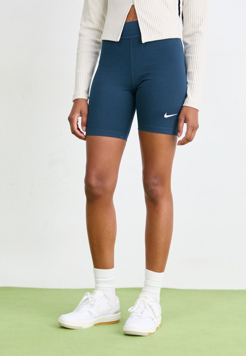 Tamsiai mėlynos Nike dviračių šortai su aukštu liemeniu, pagaminti iš elastingos medžiagos, derinami su baltomis sportbačiais ir raukšlėta trumpa striuke.