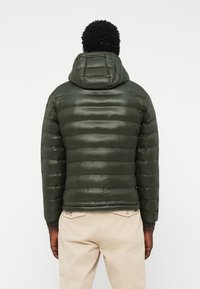 Puffer jacket in dark green with a glossy finish, featuring a hood, horizontal quilted design, and elastic cuffs. Worn with beige pants.