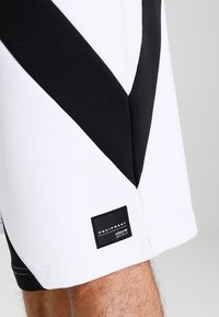 White athletic shorts featuring black geometric paneling, a textured finish, and a black label with logo on the hem.