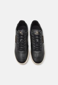 Zapatillas de cuero negro con una textura suave, suela de goma color crema, puntera redonda, cordones negros y detalles en dorado en los laterales.