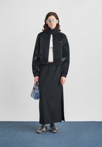 Άτομο που φοράει μαύρο τζάκετ Adidas, φούστα, λευκό μπλουζάκι, γκρι αθλητικά παπούτσια, γυαλιά ηλίου και κρατάει μια μπλε τσάντα, στέκεται σε μπλε χαλί.