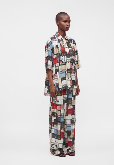 Personne portant une chemise oversize assortie et un pantalon large avec des motifs d’étiquettes rectangulaires colorées et des sandales noires à bouts ouverts, debout de côté.