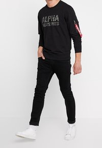 Hombre vestido con sudadera negra de "Alpha Industries", jeans negros, zapatillas blancas, y con una etiqueta roja en el bolsillo de la manga izquierda.