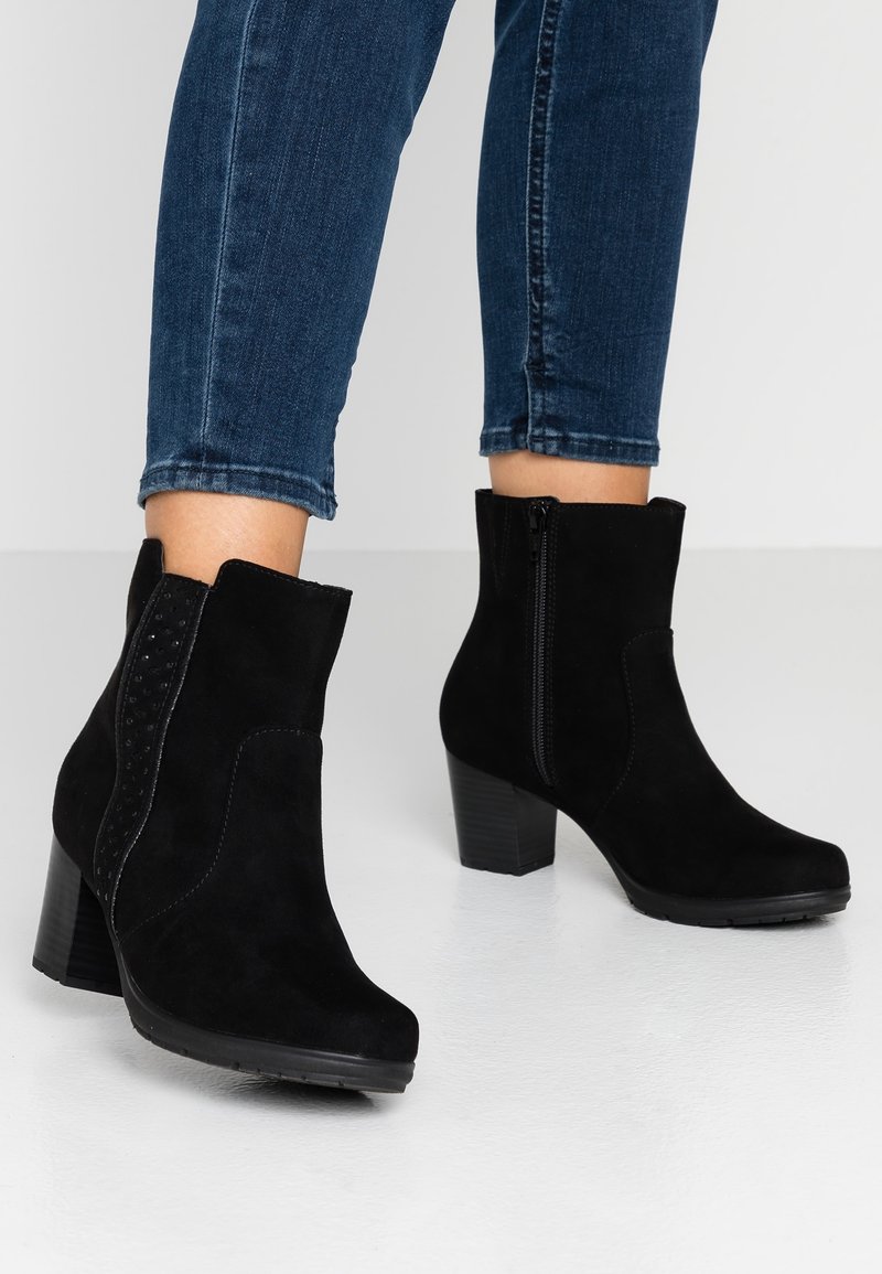 Personne portant des bottines en daim noir à talons carrés et fermetures éclair latérales, associées à un jean en denim bleu sur un fond uni.