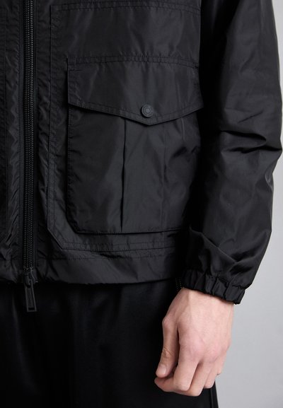 Schwarze Nylonjacke mit Reißverschluss, elastischen Bündchen und einer Klapptasche auf der Brust. Strukturierte Oberfläche mit glatter Verarbeitung.