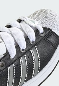 Black leather sneaker featuring white laces and three silver stripes. The toe box has a textured, white knit fabric. Circular eyelets and logo detail visible.