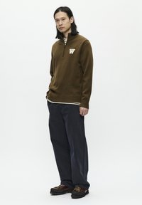 Brown knit sweater with a zip collar featuring a cream "W" logo, layered over a striped shirt. Worn with dark, loose-fitting pants and brown shoes.