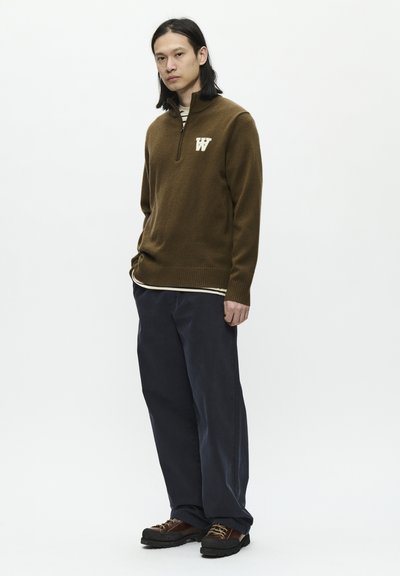 Un pull en tricot marron avec un col zippé arborant un logo "W" crème, superposé à une chemise rayée. Porté avec un pantalon large et sombre et des chaussures marron.