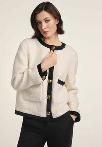 Biała, teksturowana kurtka z czarnymi wykończeniami, z dużymi złotymi guzikami i dwiema przednimi kieszeniami. Projekt zawiera okrągły dekold i luźny krój.