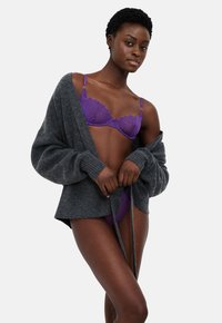 Sieviete, tērpta violetā mežģīņu veļā un brīvā pelēkā kardiganā pret vienkāršu gaišu fona, pielāgo kardigana saišu.