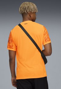 Maglietta sportiva arancione brillante con maniche a fantasia, maniche corte e etichetta sul retro che recita "MILANO." Cinghia diagonale nera attraversa il corpo.