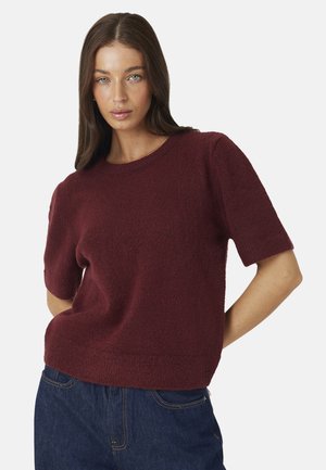 Donna con capelli castani lunghi che indossa un maglione bordeaux a maniche corte e jeans blu, in piedi con le mani dietro la schiena.