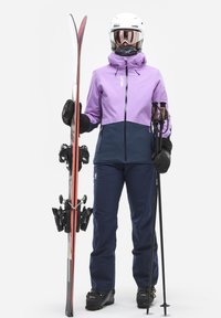 L'équipement de ski comprend une veste violette et marine, des gants noirs et un pantalon marine. Le skieur tient des skis noirs avec des accents rouges et des bâtons.