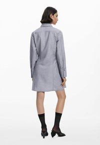 Abito camicia grigio a maniche lunghe con colletto a bottoni e spacchi laterali. Presenta una texture sottile e una vestibilità sagomata, che termina a metà coscia.