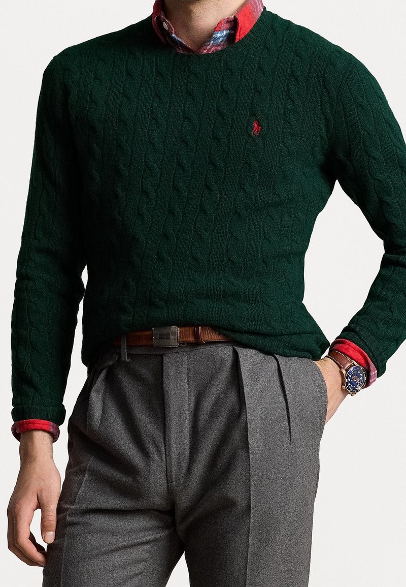 Dunkelgrüner Kabelstrickpullover mit V-Ausschnitt. Getragen über einem rotkarierten Hemd, kombiniert mit grauen, gefalteten Hose und einem braunen Gürtel.