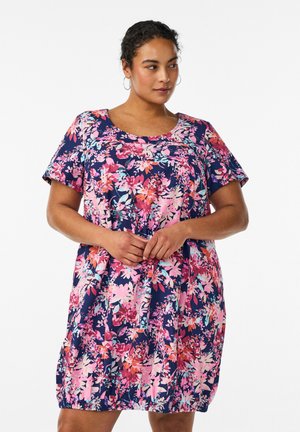 Žena v krátkom rukáve, šaty do kolien s výrazným ružovo-fialovým kvetinovým vzorom, stojaca so spojeními rúk voľne pred sebou.