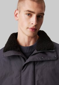 Mørk grå jakke med en fluffy svart krage og en frontglidelås. Modellen har på seg en mørk ribbet genser under.