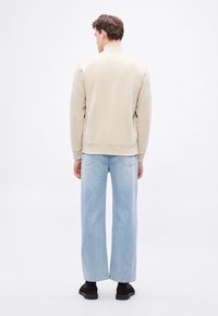 Világos bézs pulóver magas nyakrésszel, bő szárú világoskék farmernadrággal párosítva. Feketék a cipők, amelyek kiegészítik a ruházatot. Hátsó nézet, egyszínű háttér.