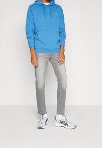 Felpa con cappuccio blu in cotone con tasca frontale e coulisse sul cappuccio, abbinata a jeans grigio chiaro e sneaker bianche con dettagli colorati.