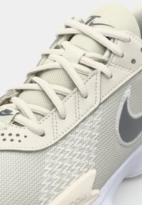 Lys grå sneakers med mesh-overdel, syntetiske overlays og polstret krave. Har flade snørebånd, teksturerede detaljer og kontrasterende sølvlogo.