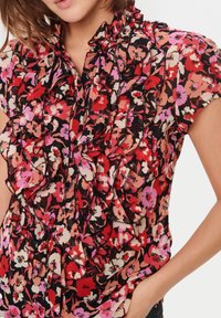 Blouse fleurie en rouge, rose et noir. Présente un col à volants et un devant avec fermeture à boutons. Manches courtes et tissu léger.