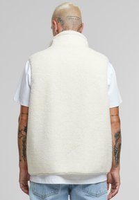 Gilet en polaire blanc avec un col haut, surface texturée, design sans manches et ourlet arrondi. Porté par-dessus un T-shirt blanc et un jean bleu.