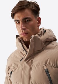 Beige polstret jakke med høj krave, lynlåslommer og hætte med snor. Glat tekstur og strømlinet design.