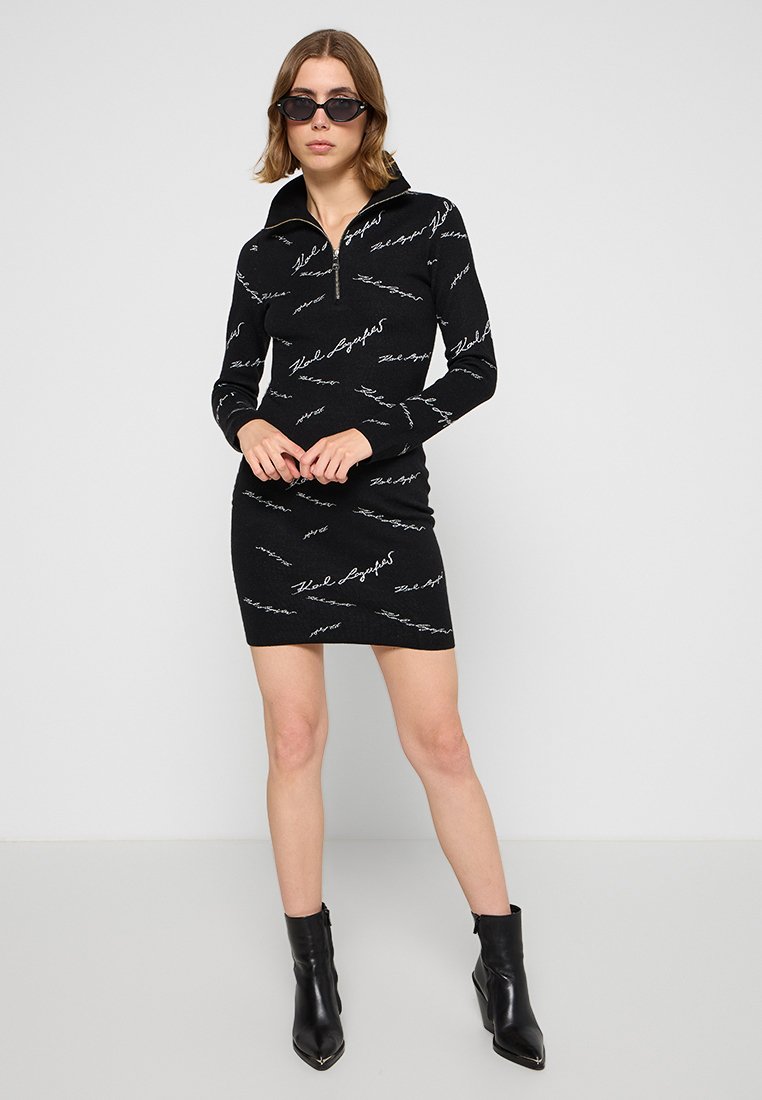 Robe noire ajustée avec des manches longues et un col zippé à moitié. Présente un motif répétitif en texte en cursive blanc. Portée avec des bottines noires.