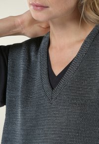Gilet gris en maille avec un col en V, tissu texturé et coupe ample porté sur un haut noir. Gros plan montrant le col et les détails du tissu.