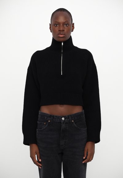 Maglione nero corto con collo alto e zip argentata, caratterizzato da una texture a coste e maniche larghe, abbinato a jeans in denim scuro.