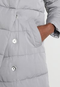 Světlě šedý prošívaný péřový kabát s prošívaným vzorem, lesklými knoflíky na zapínání, boční kapsou a hladkou texturou.