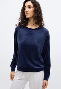 Γυναίκα με σγουρά σκουρόχρωμα μαλλιά που φορά μπλε βελούδινο μπλουζάκι με μακριά μανίκια και ανοιχτό γκρι παντελόνι, κοιτάζει προς το πλάι μπροστά σε ένα απλό φόντο.