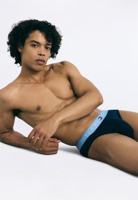Námorné pánske slipy s bledomodrým pásom, na ktorom je nápis "TOMMY HILFIGER". Model leží bez trička na bielom pozadí.