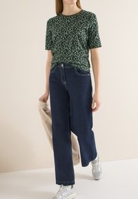 Femme portant une chemise à manches courtes verte à motifs, un jean bleu foncé à jambes larges, des baskets argentées, tenant une veste beige texturée dans la main gauche.