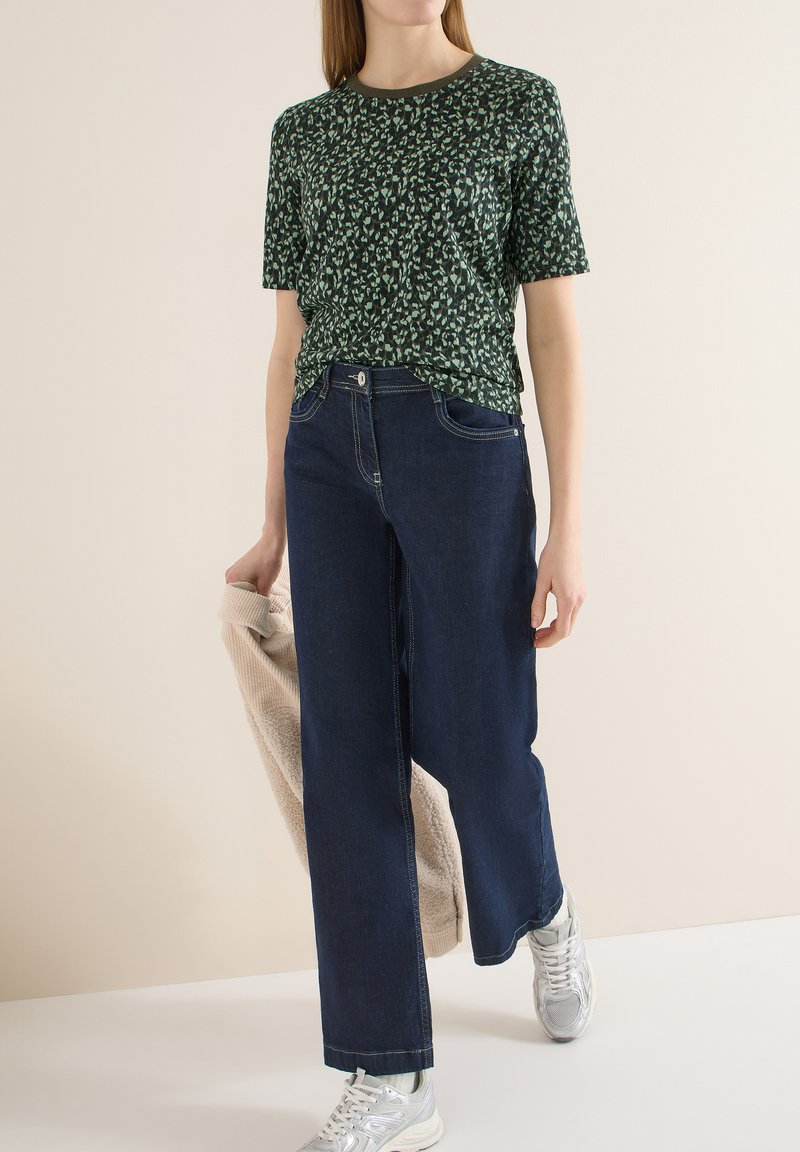 Femme portant une chemise à manches courtes verte à motifs, un jean bleu foncé à jambes larges, des baskets argentées, tenant une veste beige texturée dans la main gauche.