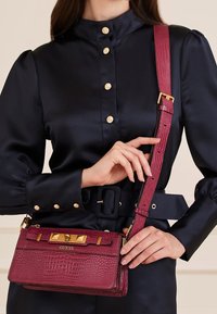 Bolso de piel texturizada color marrón con herrajes dorados; presenta una correa larga y ajustable y un diseño de cierre elaborado.