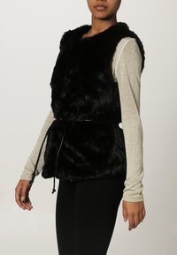 Musta tekoturkisliivi, jossa on pyöreä pääntie, vyötäröllä vyö ja pehmeä pinta, yhdistettynä kimaltavaan pitkähihaiseen toppiin.