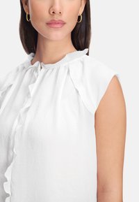 White sleeveless blouse with ruffled neckline and shoulder seams, made of light, textured fabric. Minimalist design, no patterns or accents.