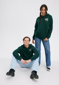 Green hoodies with front pockets, loose fit. Male model in light jeans and black boots; female model in blue jeans and black sneakers.