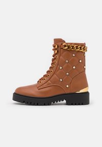 Bottine en cuir marron matelassé avec accent de chaîne dorée, clous ronds en métal, col rembourré et semelle en caoutchouc épaisse. Talon noir avec détail métallique.