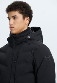 Giacca a vento nera con collo alto, cappuccio regolabile con cordino e zip. Tessuto testurizzato con dettagli del logo su manica e scollatura.