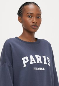 Felpa blu navy con scritte bianche "PARIS" e "FRANCE". Presenta una scollatura rotonda e una vestibilità morbida con tessuto dalla texture soffice.